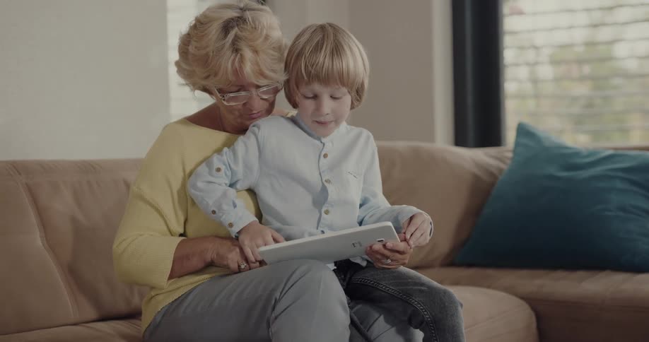 A boy plays with a computer tablet while sitting on the lap of an elderly woman in the living room of a house during the day