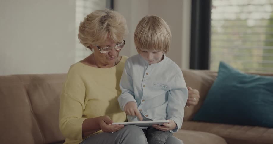 An elderly woman with blond hair and glasses is sitting on the couch when a little boy brings a tablet and sits on her lap