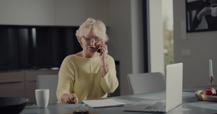 A woman is sitting at a kitchen table and talking on her cell phone