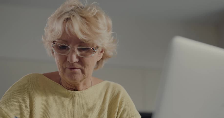 Elderly woman with fair hair glasses and yellow shirt works on laptop