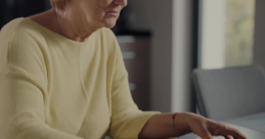 A woman using a laptop at a table writes notes in a spiral notebook
