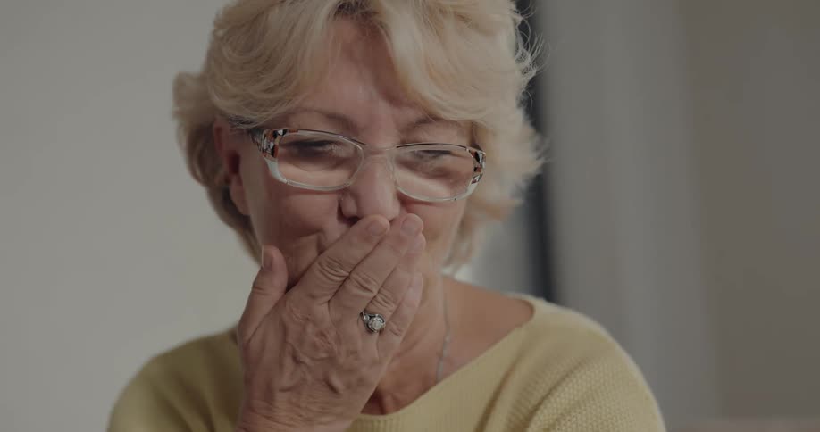 Elderly woman with fair hair glasses necklace and yellow sweater makes video call on smartphone waving and smiling