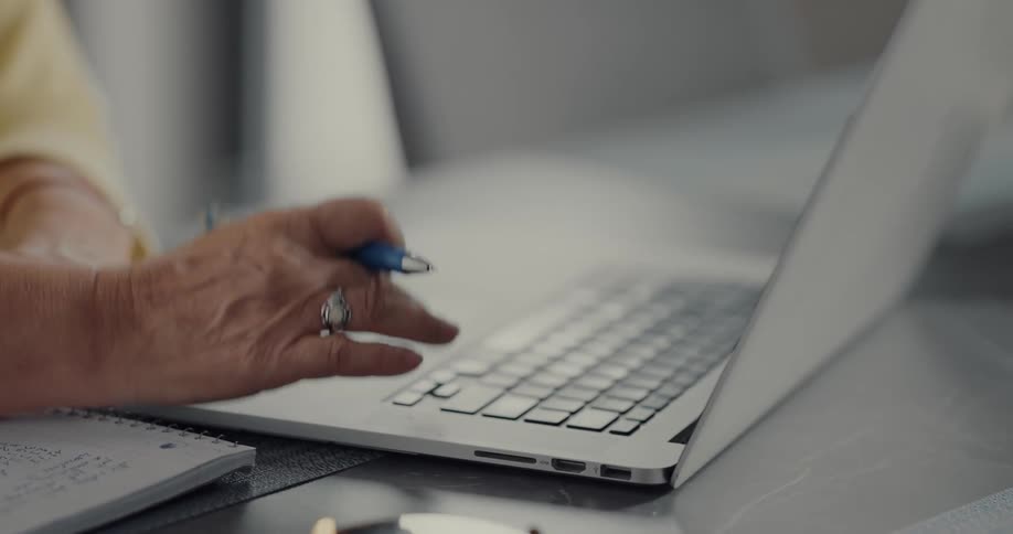 Female senior citizen writes notes with pen and paper while using a laptop