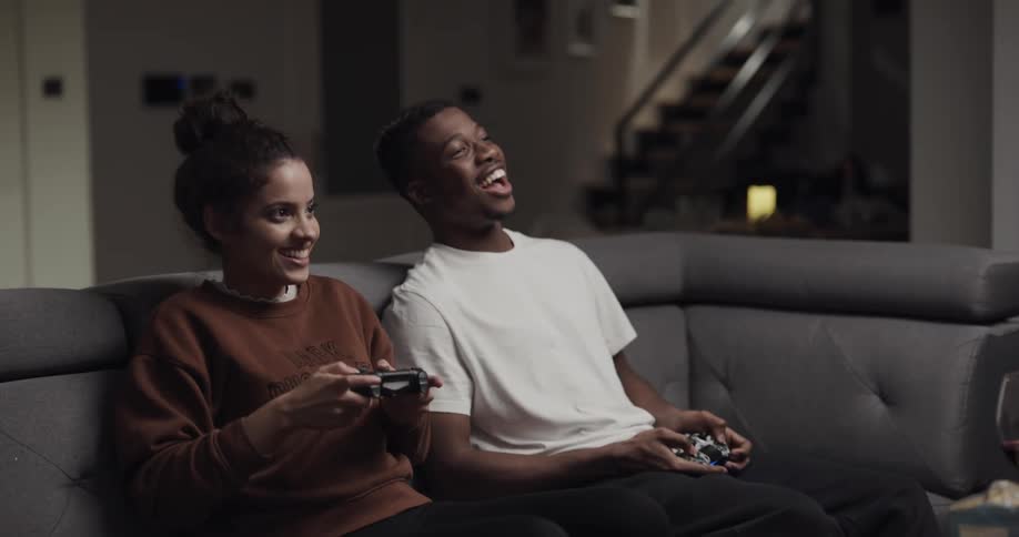 A man tries to obstruct the woman playing video games against him as they sit together on a sofa in the living room of a house