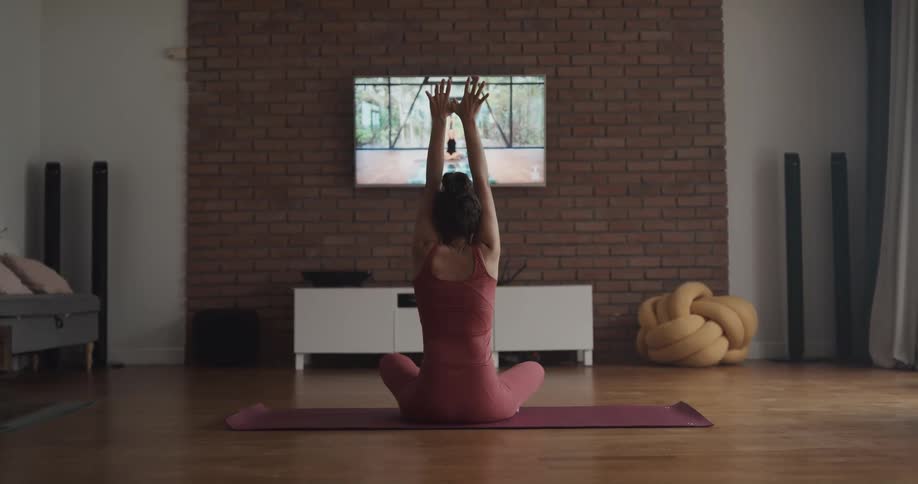 A woman is doing stretches while watching a yoga video on a tv