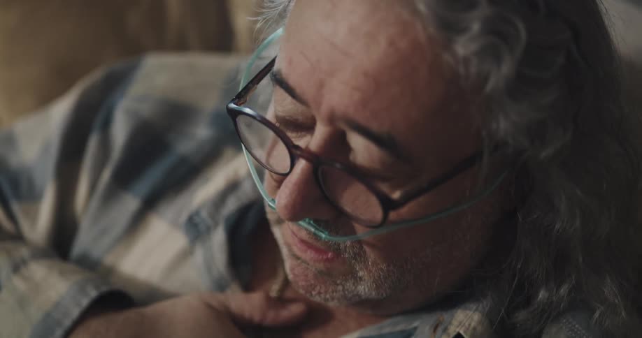 An elderly man with an oxygen tube sits on a couch coughing