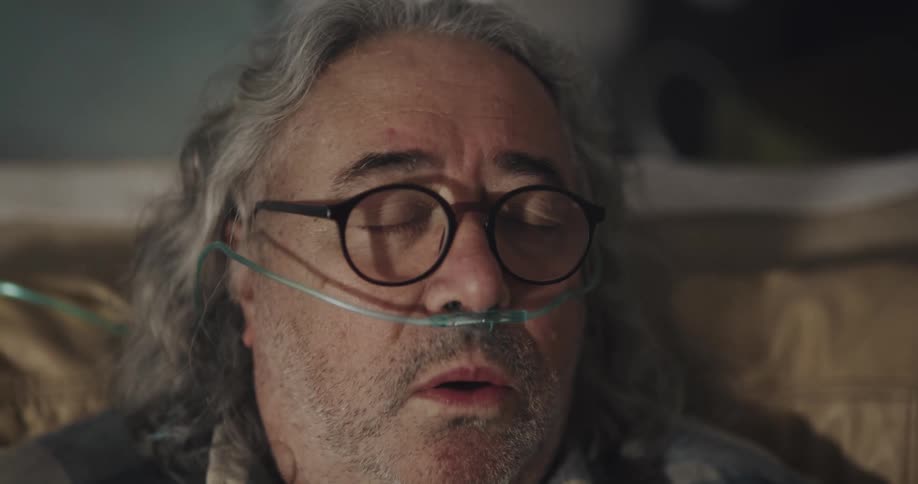 Mature man with long curled hair wearing a nasal canula has a coughing fit at home