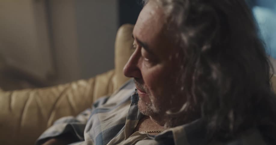 Man with gray long hair and plaid shirt sits on brown sofa drinking alcohol in living room