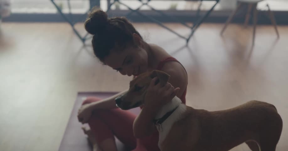 A woman is sitting on a yoga mat in her living room and petting a dog