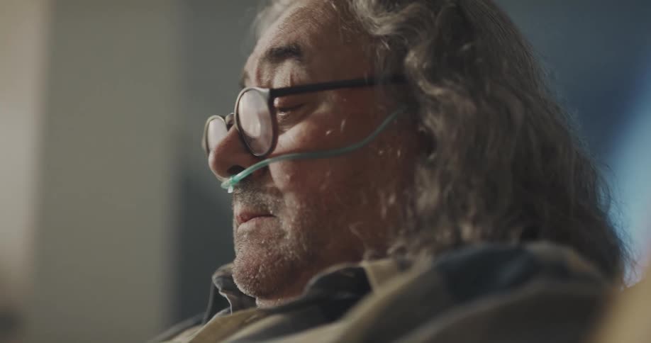 Mature man with long hair coughs while wearing a nasal cannula while sitting at home