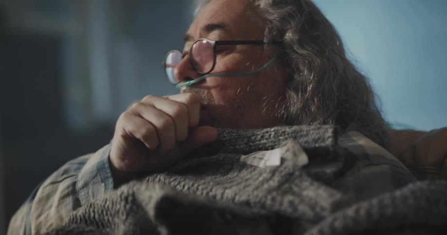 A man is sitting in a chair indoors and coughing