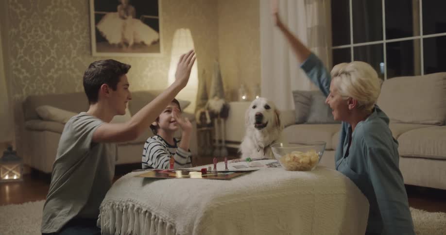 A family congratulate one another with high fives as they play a board game together in the living room of a house at night