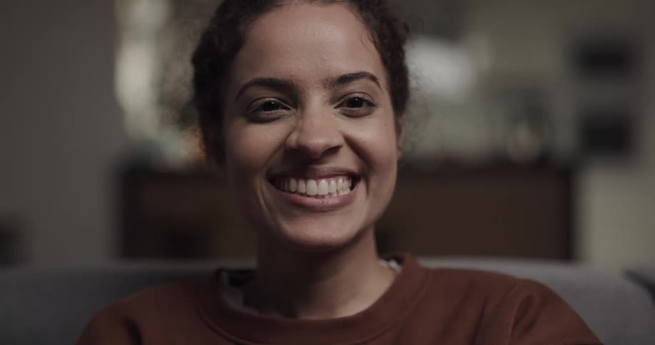 A young woman raises her gaze to smile into the camera while sitting on a sofa indoors