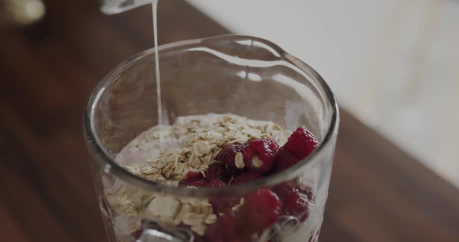 Woman pours milk over berries and oats inside a mixer in the kitchen at home
