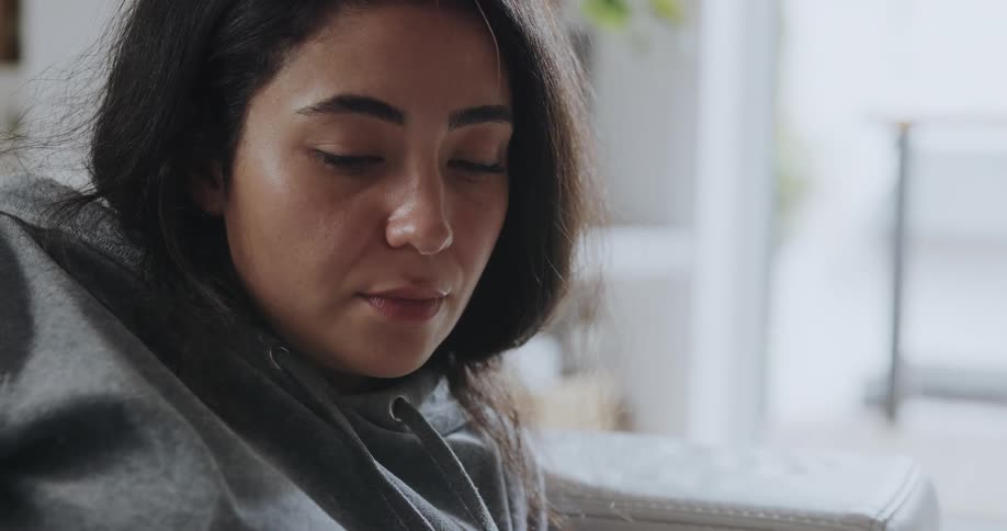 Woman with dark hair and gray hoodie lies on sofa drinking from cup in living room at home
