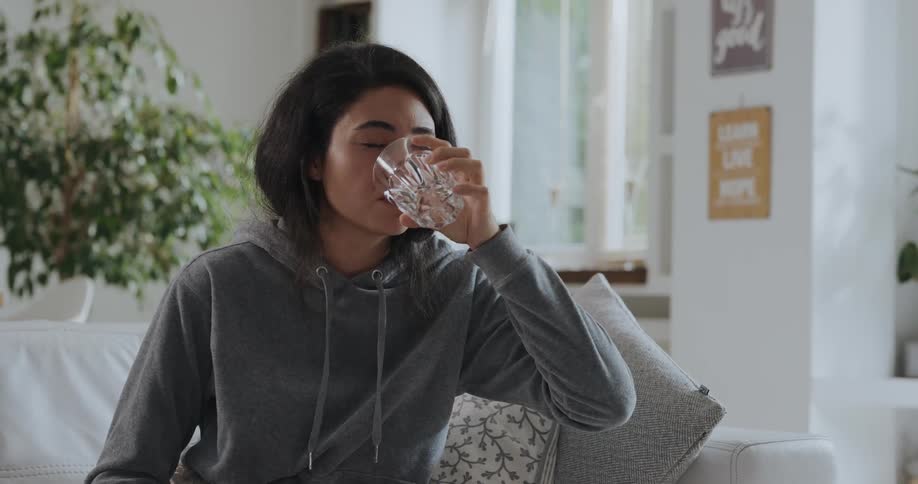 An ill woman removes a pill from its packaging and swallows it with a sip of water before lying down to rest on a sofa
