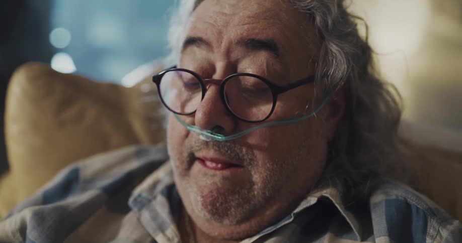 An elderly man with a breathing tube sits at home and coughs