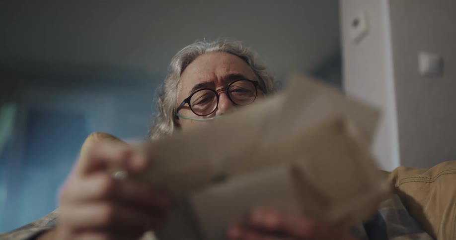 Mature man wearing a nasal canula reads old letters while sitting in the couch
