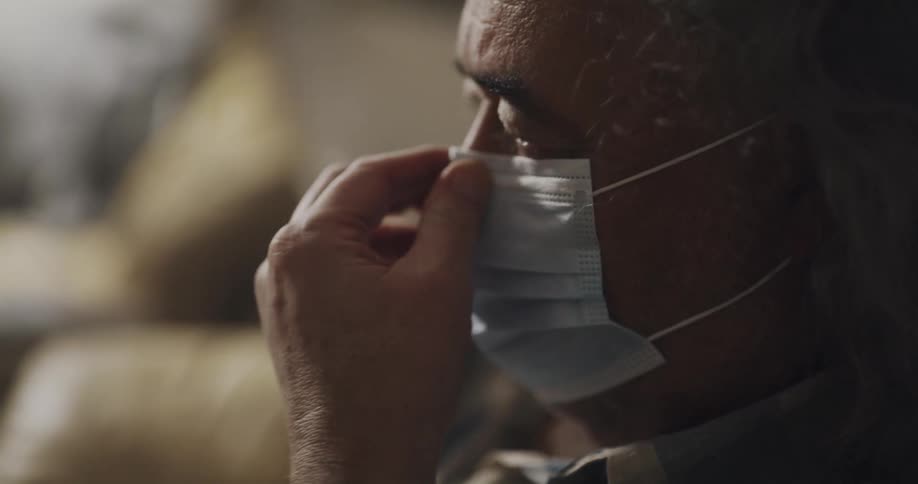 A man is sitting indoors and putting on a face mask