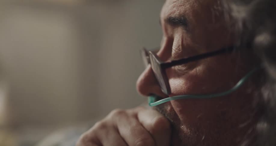 A man with a breathing tube sits at home and coughs