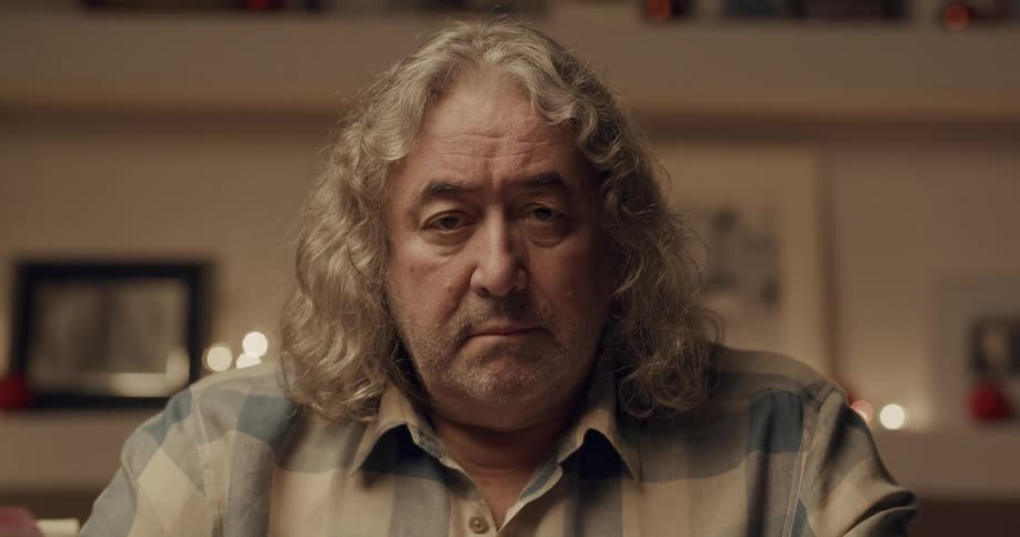 A man with long hair and unshaven face sitting in front of a wall covered with framed pictures glances up and gazes into the camera while pursing his lips slightly