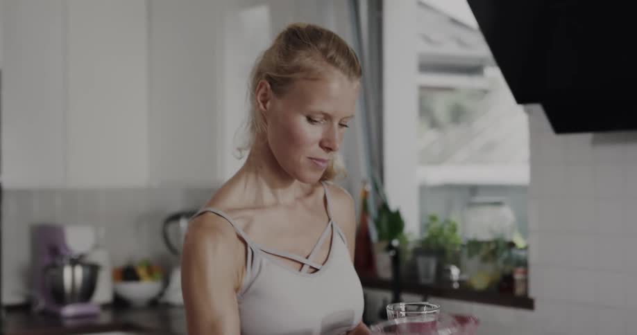A woman pours a blended smoothie from a blender into a drinking glass and takes a drink while standing in a home kitchen during the day