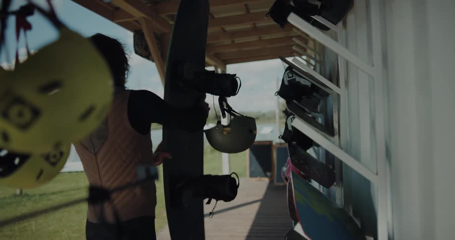 Young man wearing a black wet suit picks up a surfboard from a rack on the side of a house
