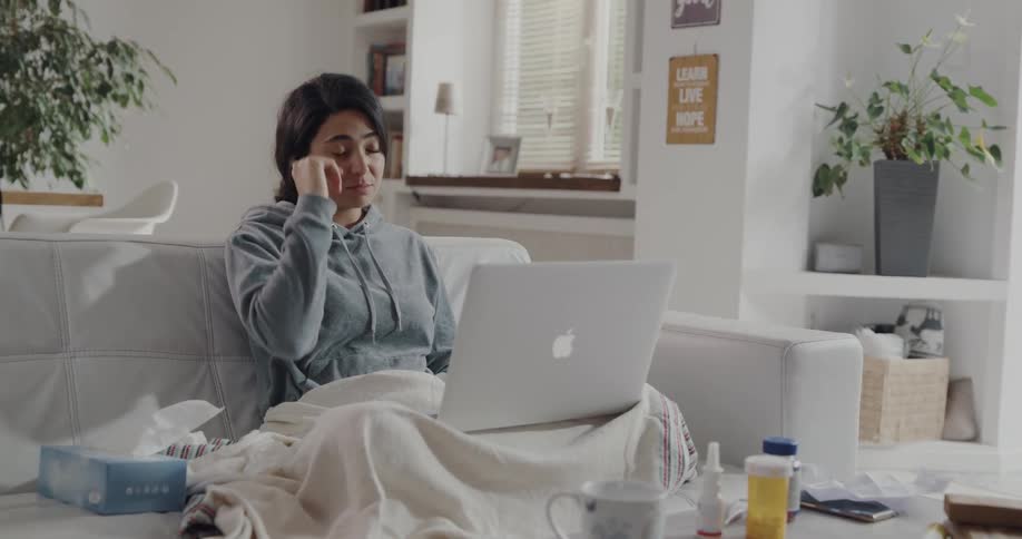 A woman sits on a couch at home and video chats on her laptop as she describes her illness symptoms