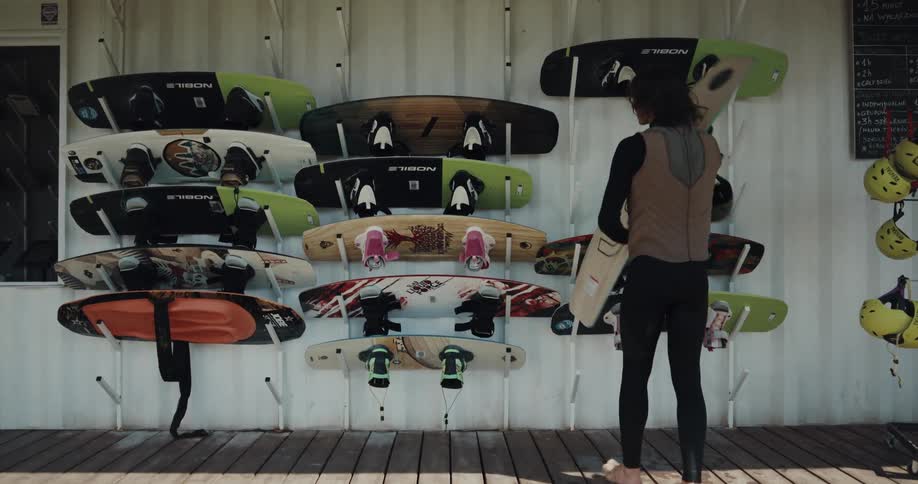 Young man wearing a black wet suit picks up a board from a rack on the wall of a house