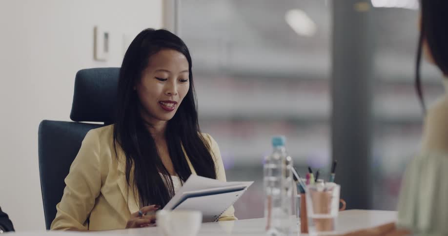 A woman in a meeting is handed a sheet of paper from another woman sitting across from her at the conference table