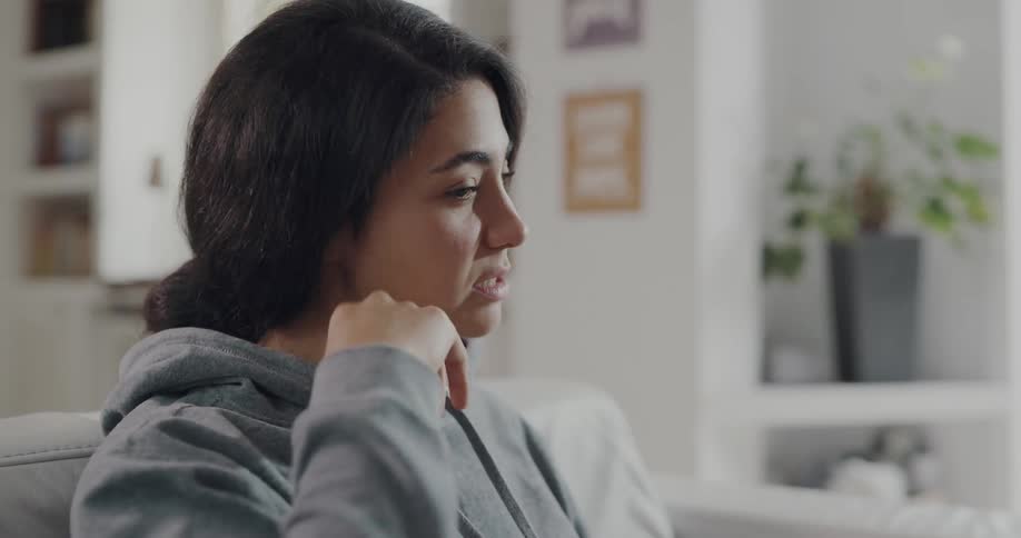 Woman with dark hair and gray hoodie sits on couch making video call on laptop in living room