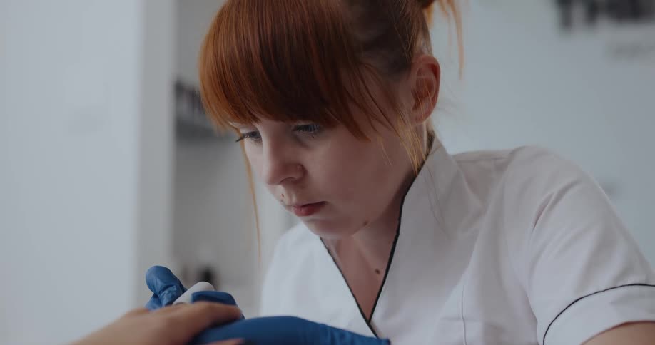 A young redhaired woman wearing blue gloves concentrates as she applies fingernail polish to a clients fingernails