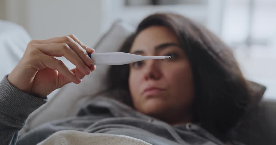 Young sick woman resting on the couch at home takes her temperature with a thermometer