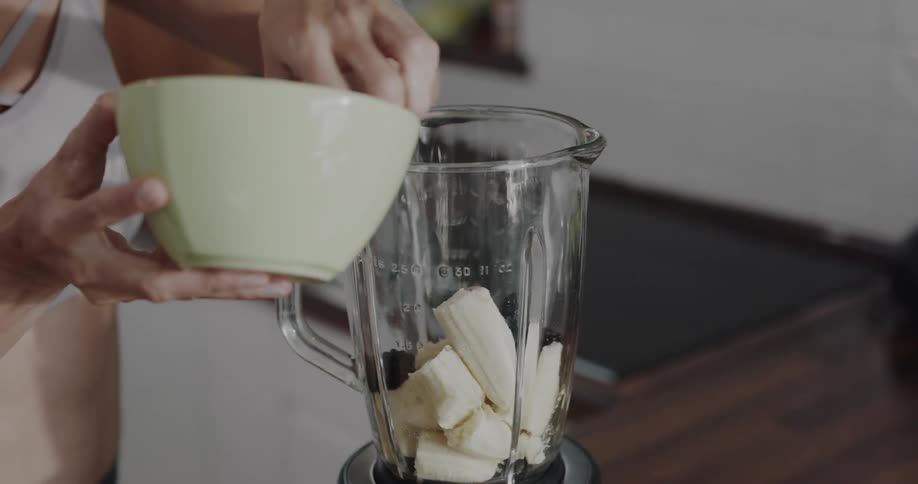 Young woman wearing sports clothes puts fruits on a blender in the kitchen at home