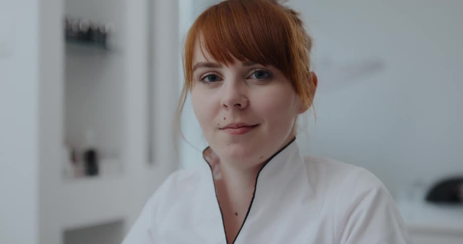 A redheaded woman is looking down blinks and then raises her head to gaze into the camera as she smiles slightly