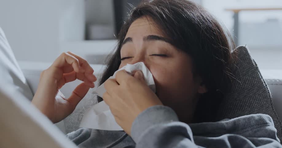 A woman using her smartphone while lying on sofa sneezes into a tissue