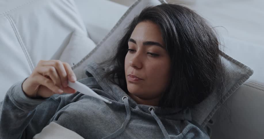 A woman resting on a sofa uses a thermometer to check her temperature before shaking her head in disappointment upon reading the measurement