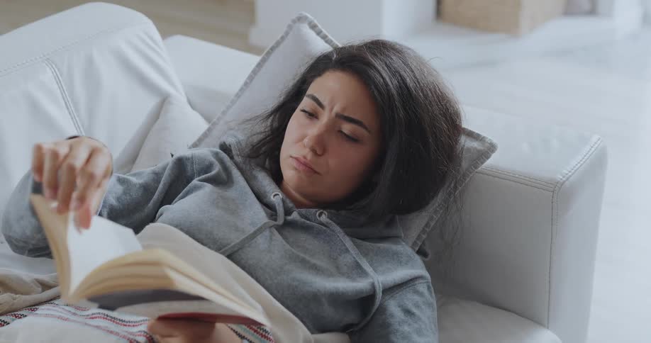 A woman is laying on a couch and reading a book