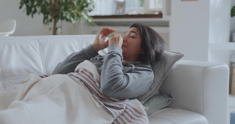 A woman is laying down on the couch and blowing her nose