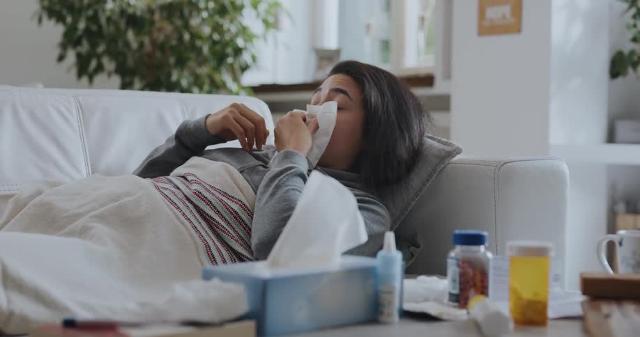 An ill woman lying on a sofa in the living room of a house sneezes into a tissue