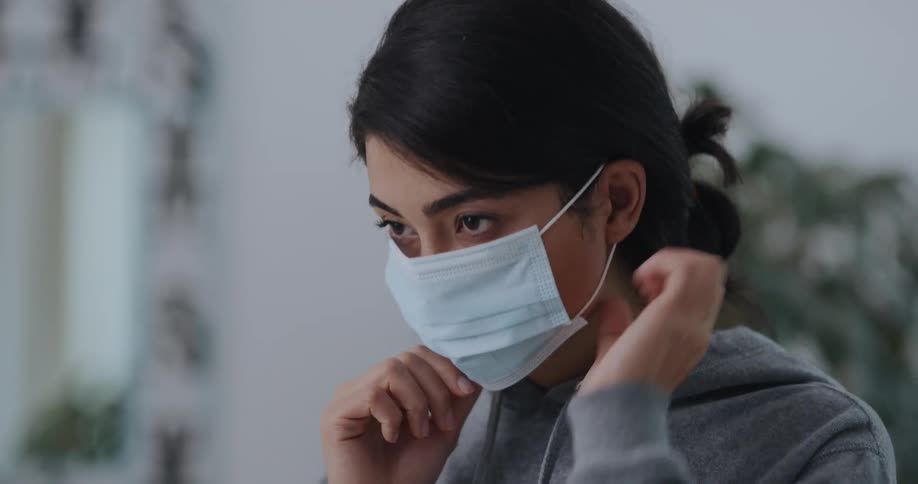 A woman takes a surgical mask from a table and puts it on her face before walking out of frame