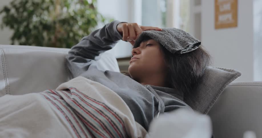 A young woman lies on her couch at home and puts a washcloth on her forehead