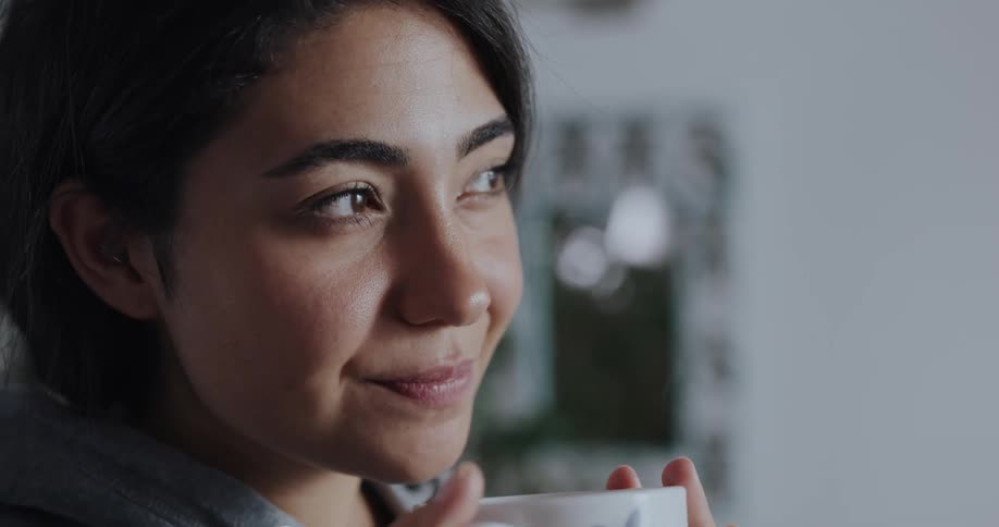 A woman clows into the cup she is holding and then sips the beverage and swallows with a contemplative look on her face