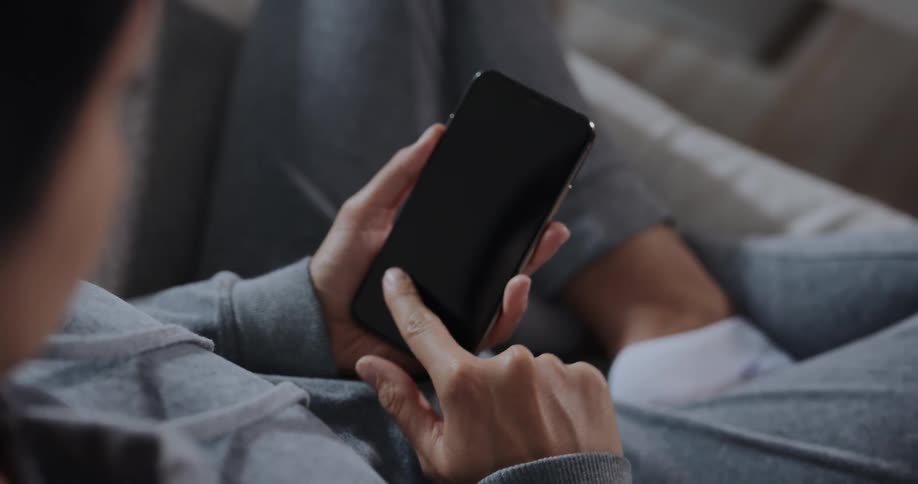 Woman with gray hoodie and pants uses smartphone sitting on couch in living room