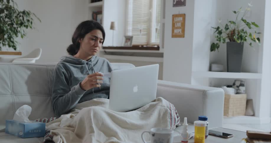 A sick woman typing on a laptop rubs neck before beginning to read a sheet of paper while blowing her nose