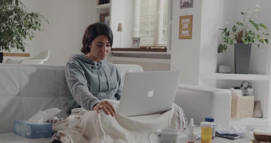 Young sick woman sitting on the couch describing her illness on a video call on a laptop