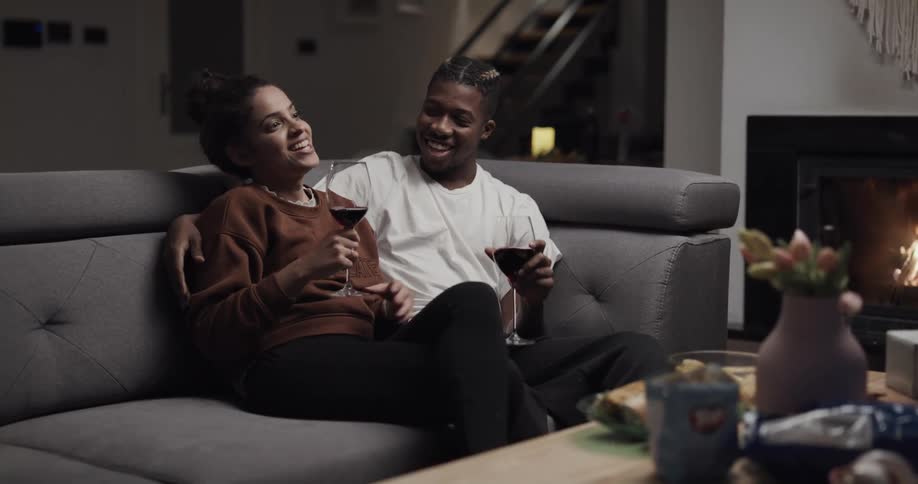 Young couple drinking wine sitting on a couch at home near a fireplace