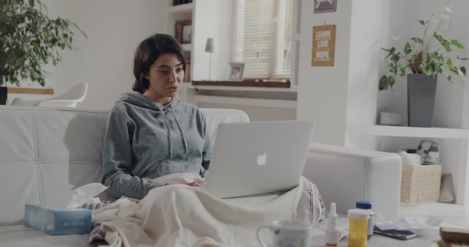 Young sick woman sitting on the couch describing her illness on a video call on a laptop