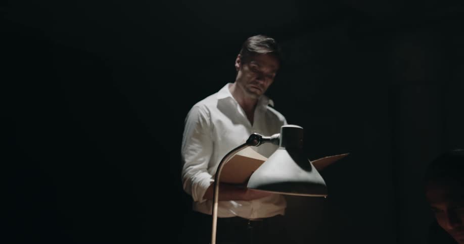 Men reading documents and having an argument inside a dimly lit room under a fan