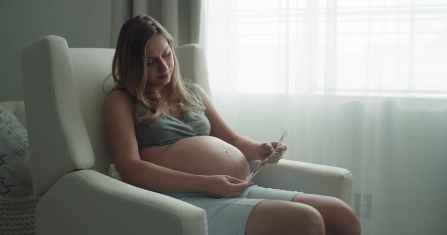 Blonde woman with green top and blue shorts strokes her belly and looks at photo sitting in armchair